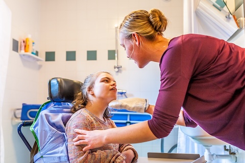 a nurse lifting up the chin of a patient with IDD, steated in a wheelchair, to look at her eyes