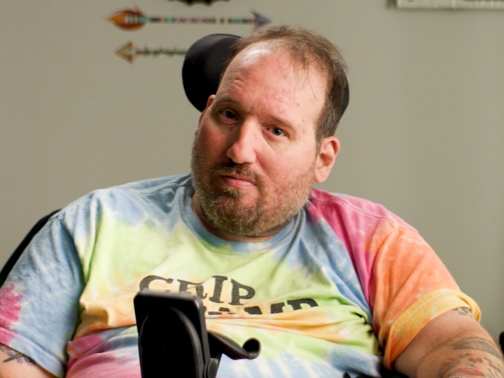 BJ Stasio sitting in a electric wheelchair wearing a tie dye shirt and smiling at the camera