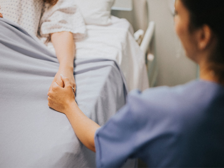 A nurse sits at a patient's bedside and holds their hand.