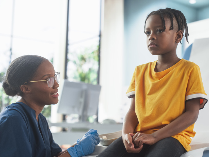 A doctor talks to a child. The doctor is squatting down, and the child is sitting on an exam table.