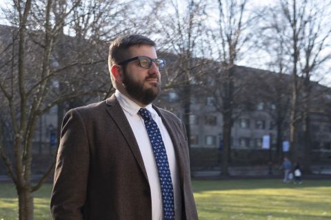 A man standing on a campus wearing a jacket and tie.