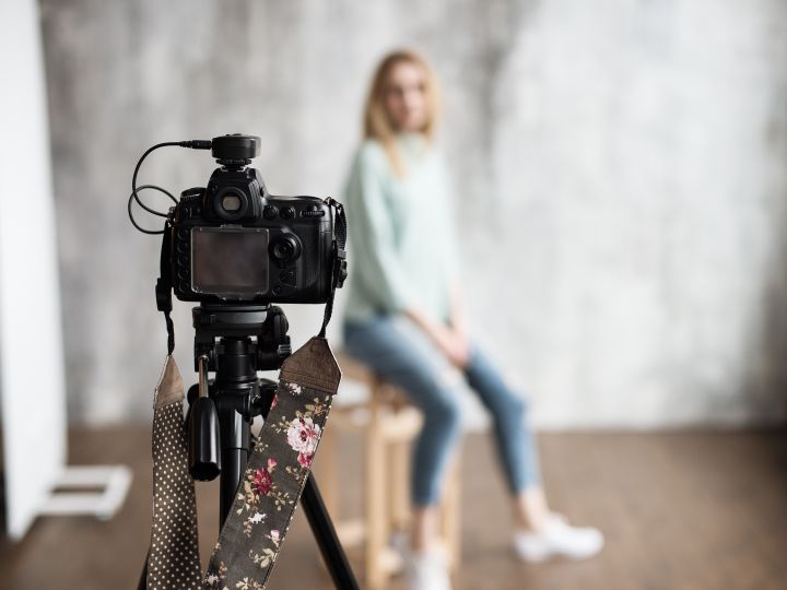 A woman sits for a photograph in a studio environment. She is in the background and blurry. The camera, on a tripod, is clear and in the foreground.