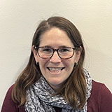 A woman with long brown hair and glasses. She is wearing a red cardigan and a patterned scarf.