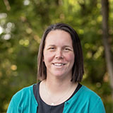 A woman with shoulder-length brown hair. She is wearing a black shirt and a teal cardigan. There is greenery behind her.