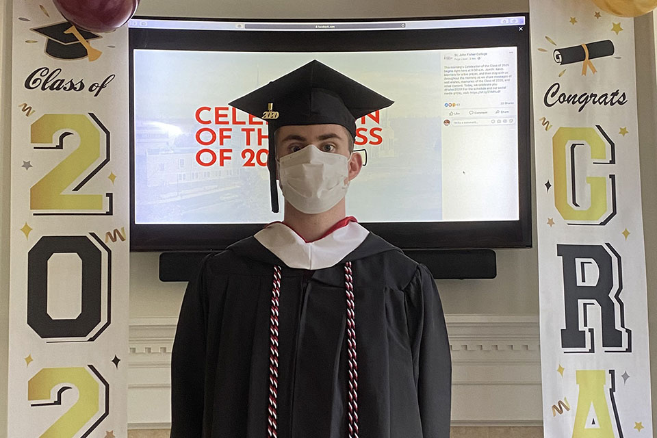 Dylan MacQuoid in cap and gown and mask in front of congratulatory banners.