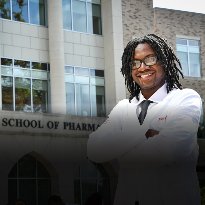 A pharmacy student stands in front of the Wegmans School of Pharmacy in her new white coat.