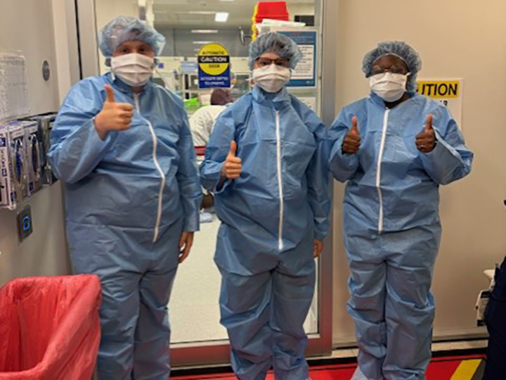 Students in protective gear and masks give thumbs up before entering a pharmacy lab.