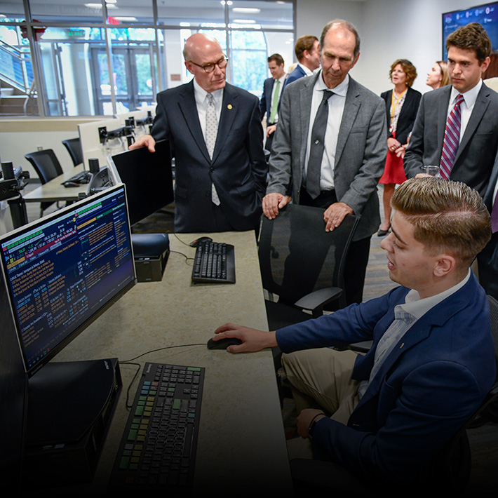 A student shows work on a Bloomberg Terminal with investors and business people standing around.