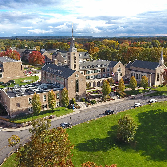 Aerial view of St. John Fisher University with colorful leaves on trees, bright green grassy hills, and sun glistening on buildings.