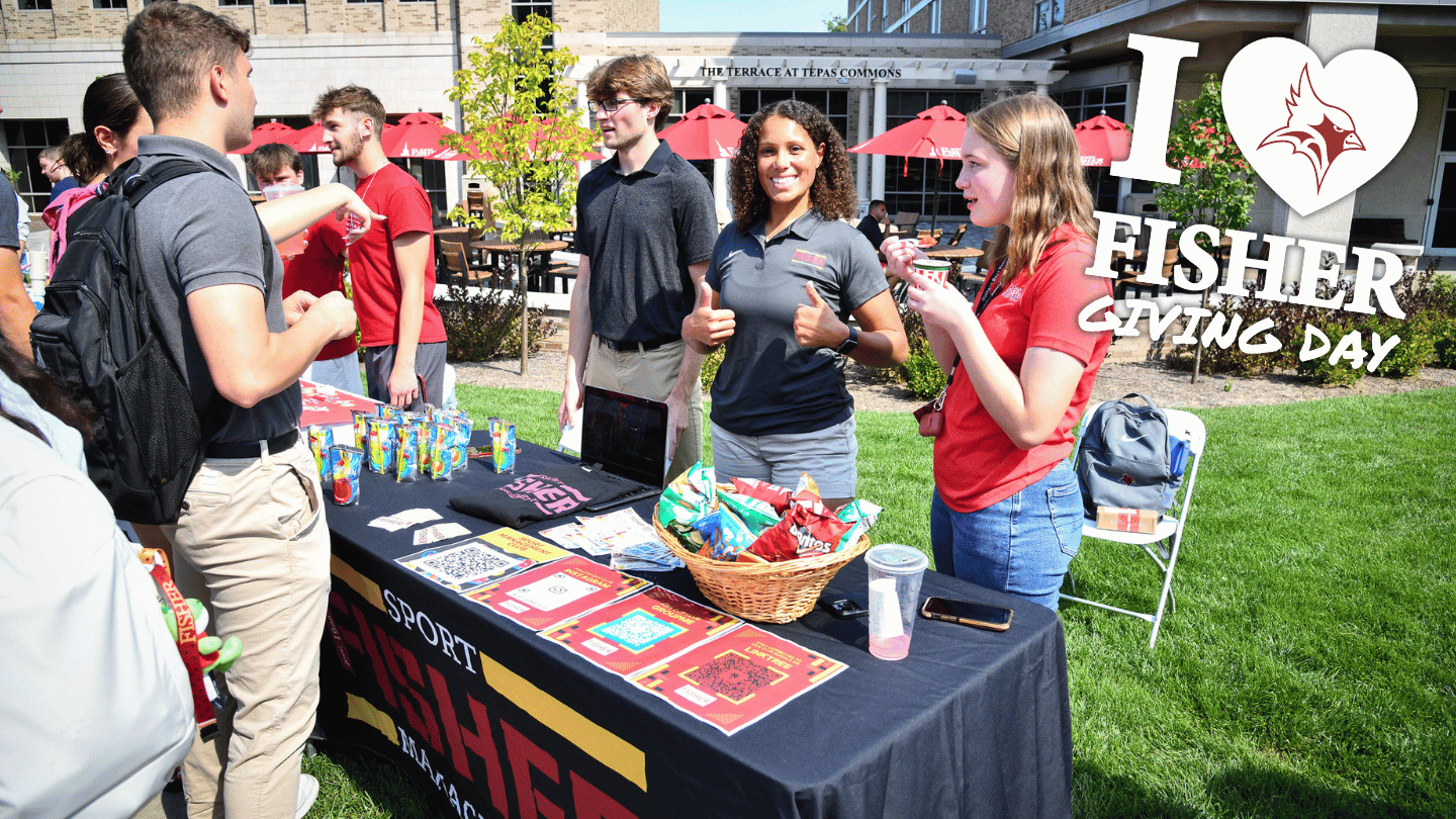 I Heart Fisher Giving Day a student gives two thumbs up while standing behind a recruitment table.