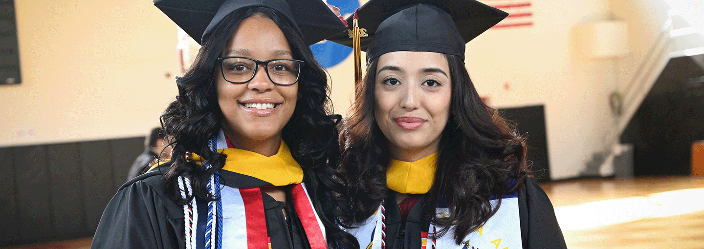 Two graduates stand together in commencement regalia.