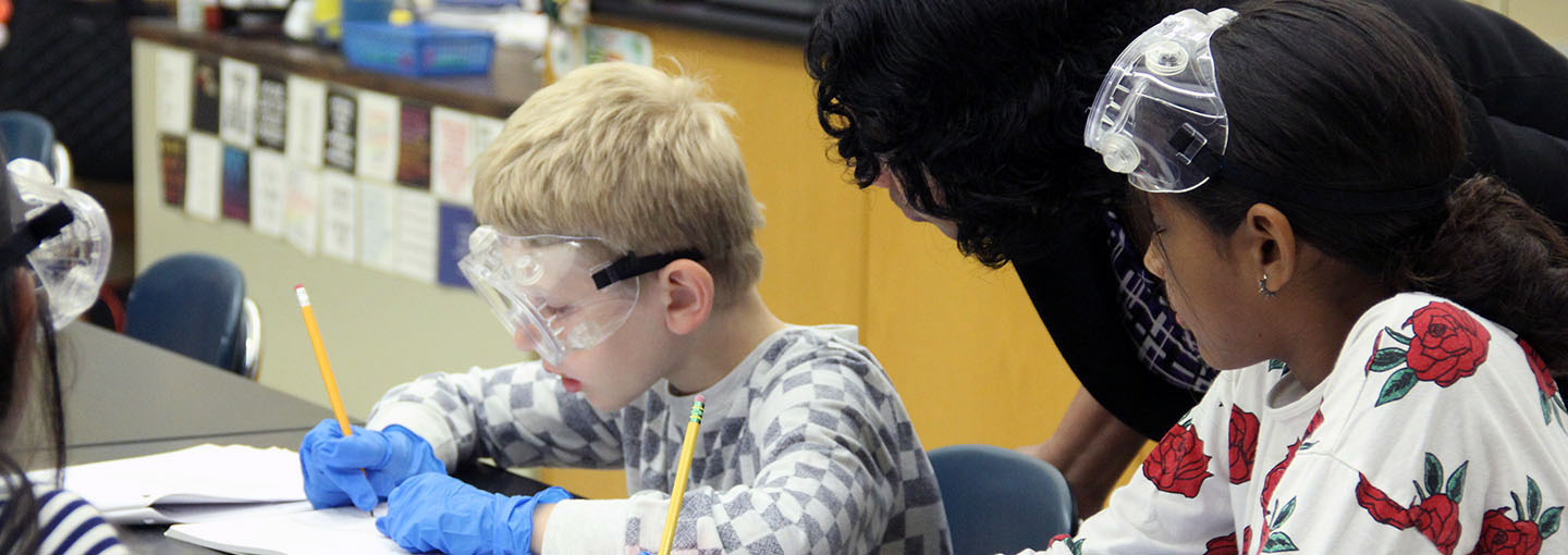 Teacher bending over to speak with two students at desks