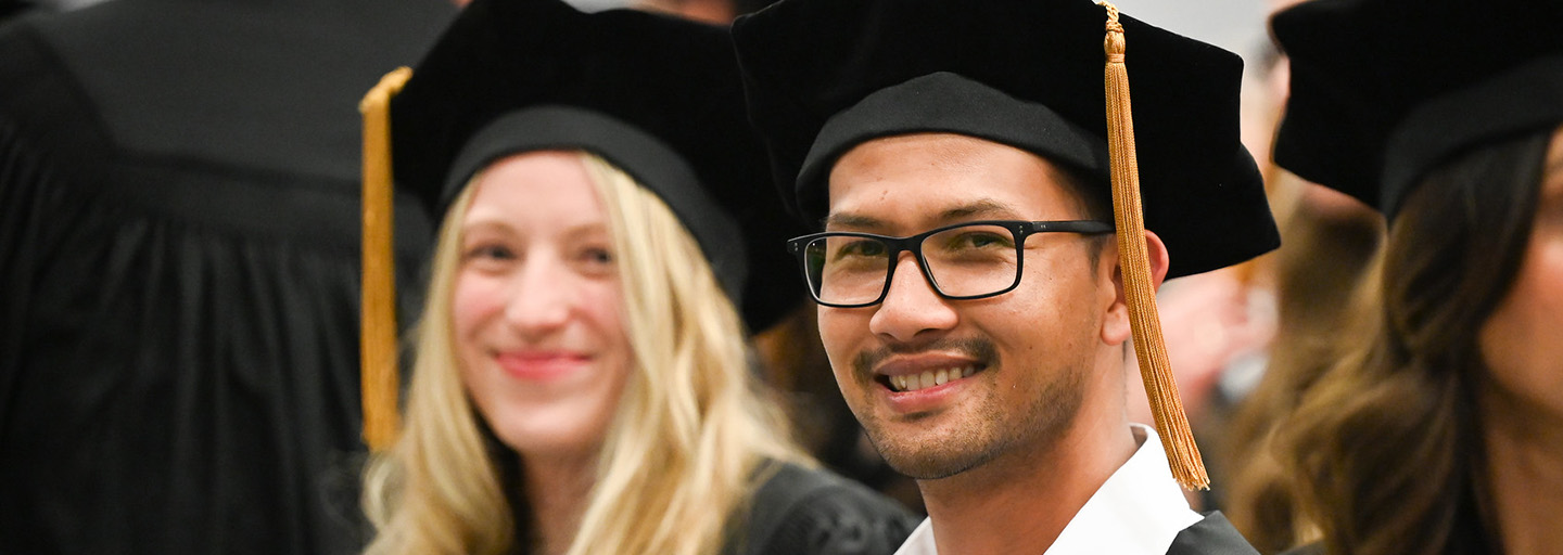 One graduate student in focus smiles, surrounded by several graduate students sitting together in commencement regalia.