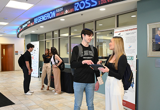 School of Business students look at information on an iPad together with a stock market ticker in the background.