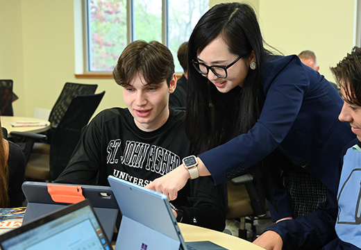 A professor points to an iPad while working with a student in a classroom.